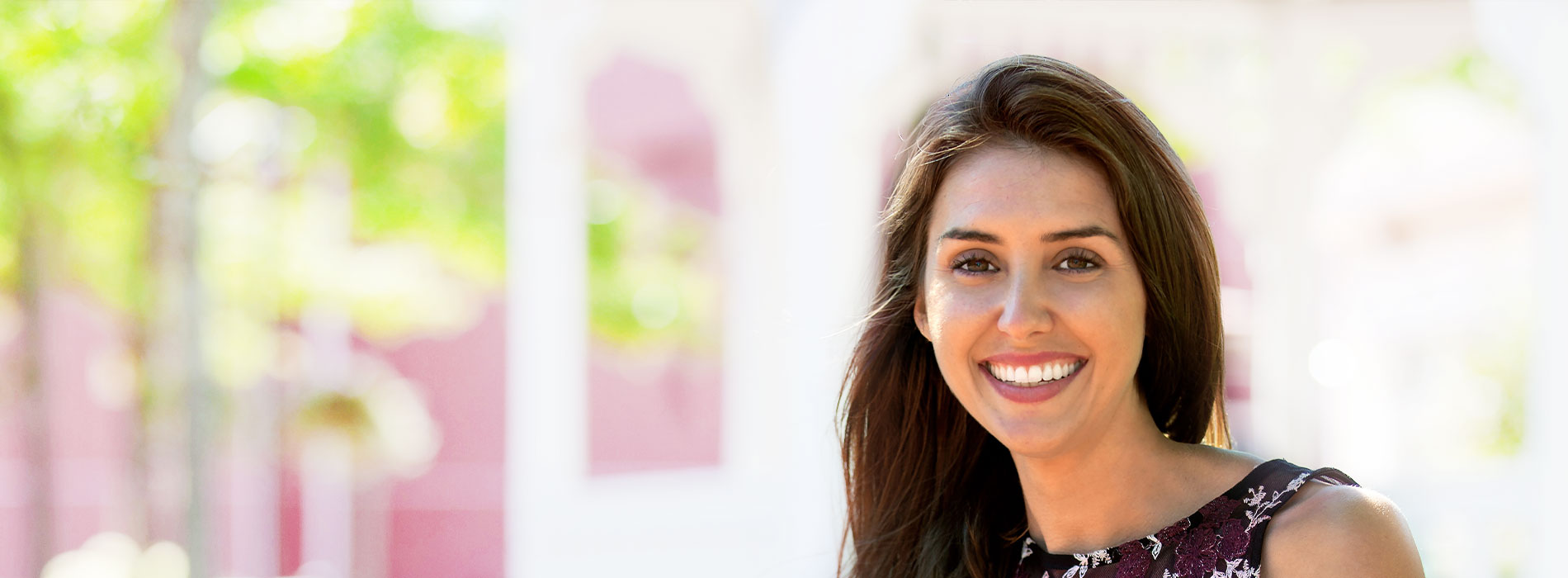 The image is a photograph of a smiling woman with long hair, wearing a sleeveless top and standing outdoors against a backdrop that includes trees and a building.