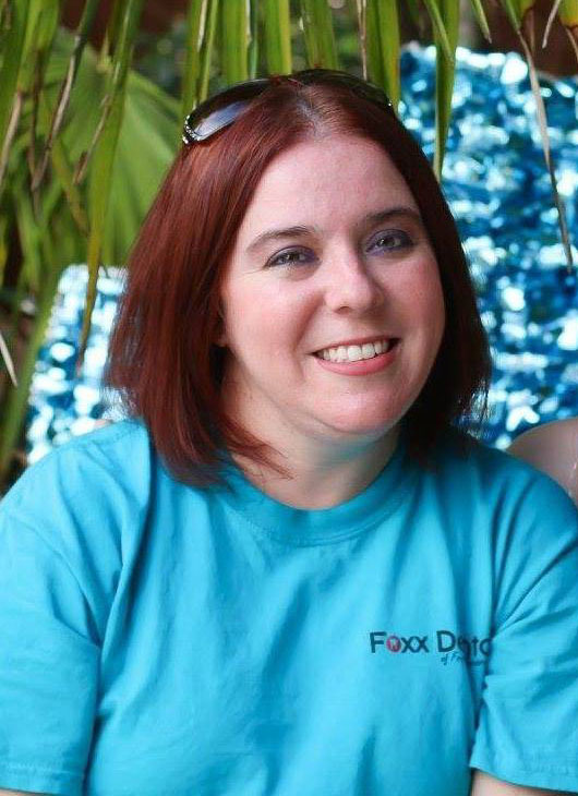 The image is a photograph of a woman with red hair and glasses, wearing a blue shirt. She has a smile on her face and appears to be outdoors, possibly at a poolside or similar location.