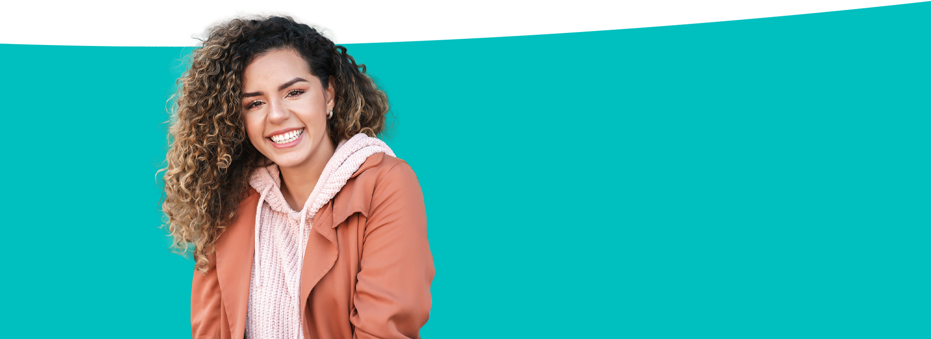 A person with curly hair is standing against a blue background, smiling and wearing a light-colored jacket.