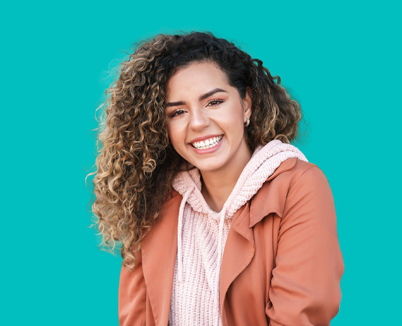 A young woman with curly hair, wearing a light tan jacket and a pink scarf, smiling and looking directly at the camera.