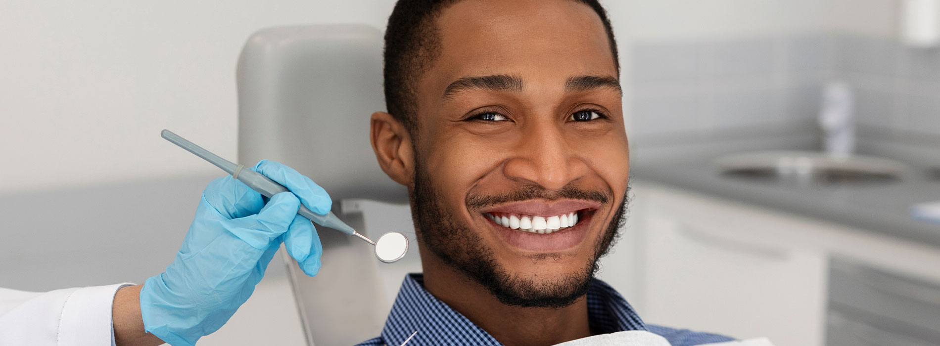A man sitting in a dental chair with a smiling expression, holding a toothbrush, and wearing a blue surgical mask.