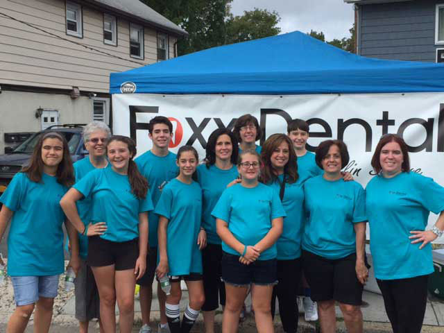 The image depicts a group of individuals, likely dental professionals, posing together outdoors in front of a tent with the word 'Foxy' on it. They are wearing matching blue shirts and appear to be participating in an event or gathering related to dentistry.