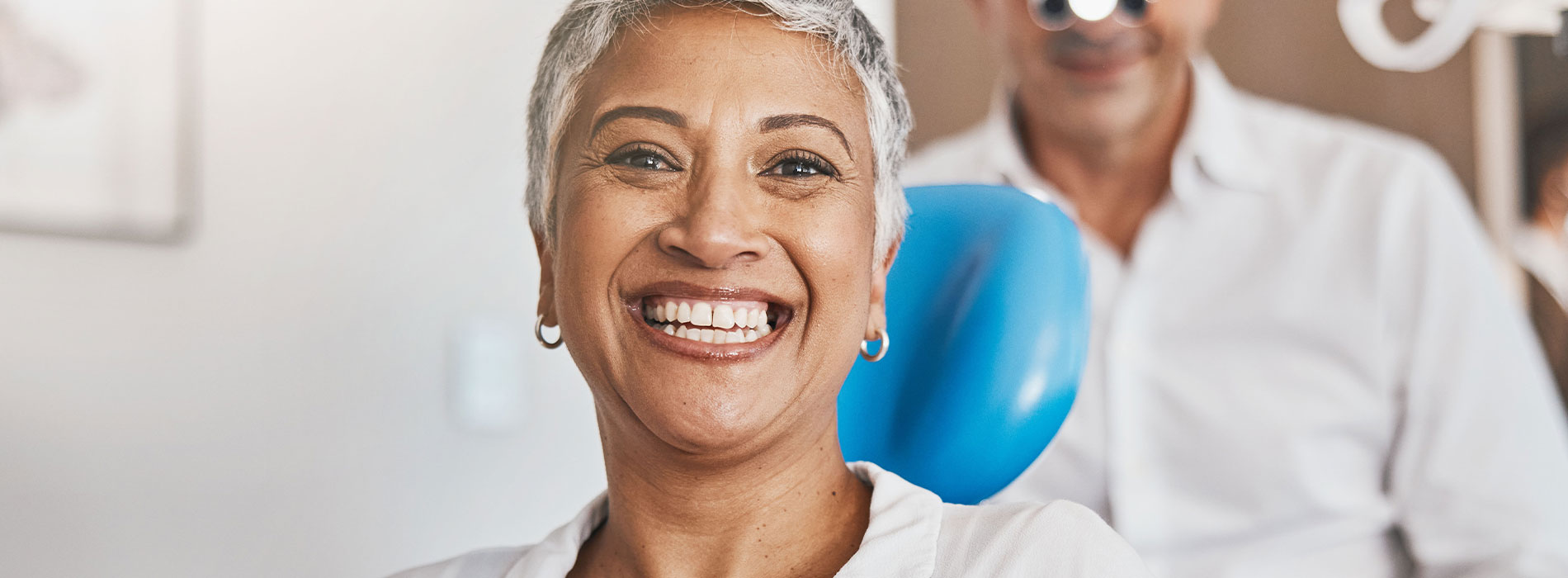 The image shows a person with a broad smile, sitting in a dental chair with a dentist behind them, both wearing masks and eye protection.