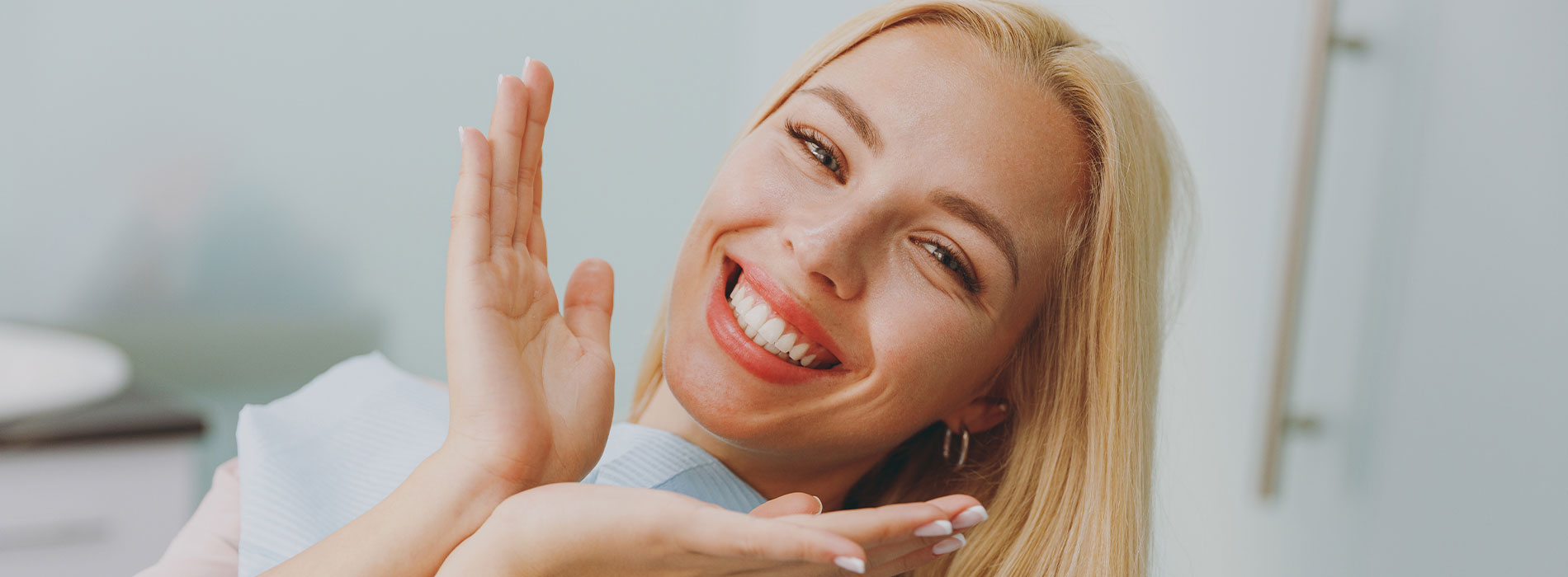 A young woman with blonde hair is smiling and waving her right hand, appearing to be in a professional or casual office setting.