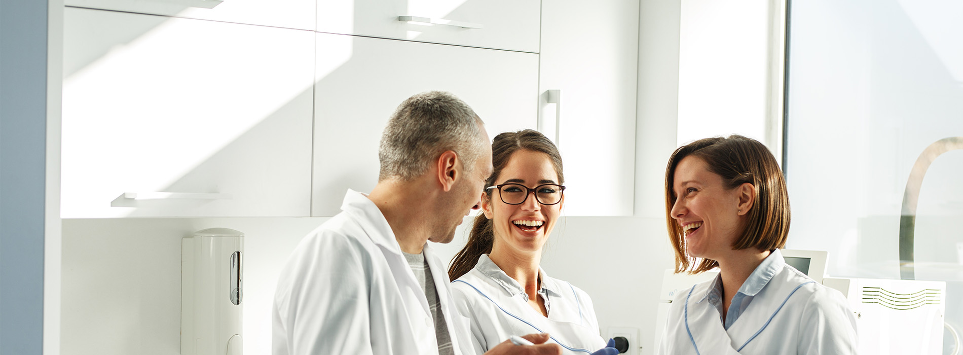 Three individuals in a professional setting, likely a dental or medical office, smiling and engaged with each other.