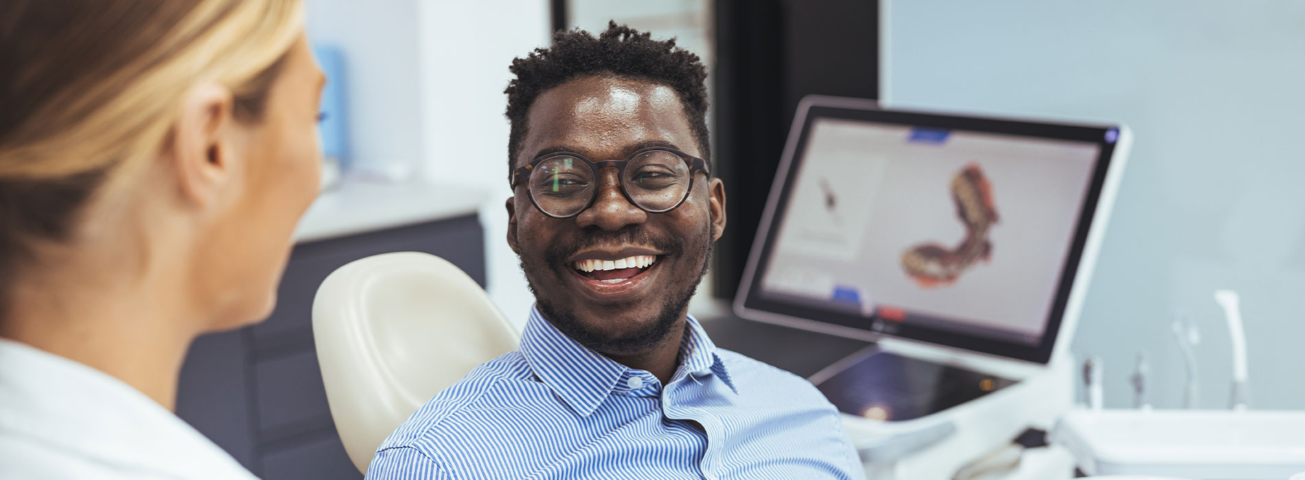 A man in a blue shirt is seated with a smile, looking at a woman who is standing and smiling behind him. Both are in an office setting with a large screen displaying medical imagery.