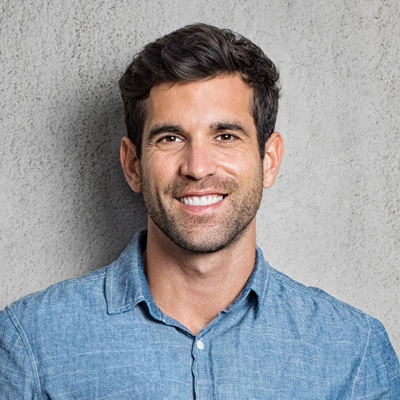 The image shows a man smiling at the camera, wearing a blue denim shirt and standing against a grey background.