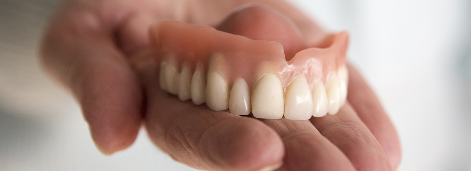 An adult hand holding a set of artificial teeth with visible dentures and a blurred background.