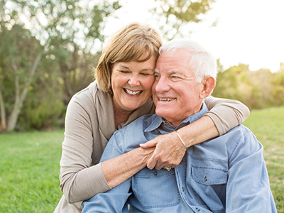 An elderly couple, a man and woman, are embracing in a warm hug. They appear to be outdoors during the daytime.