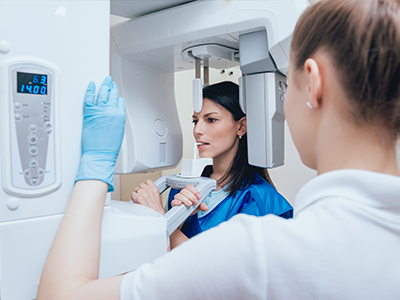 A woman in a blue coat stands next to a large, modern CT scanner, while another person looks on from behind the machine.