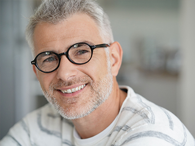 The image is of a man with gray hair, wearing glasses and a white shirt. He has a beard and mustache, and is smiling at the camera. There's no text on the image.