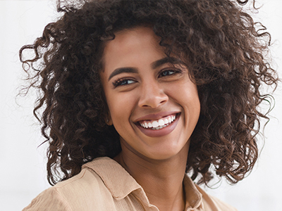 The image shows a person with curly hair smiling at the camera, wearing a light-colored top and looking directly into the lens.